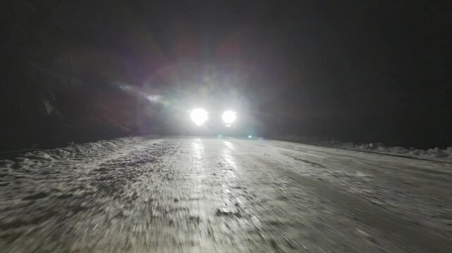 Pickup truck is driving down a snowy road at night. The headlights are on, illuminating the road ahead.