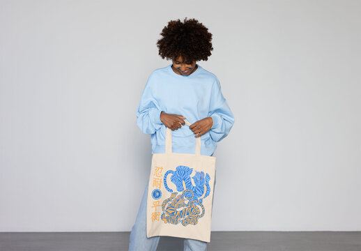Light-colored tote bag held by afro black woman in a blue sweater, great for custom logos and perfect for any occasion.