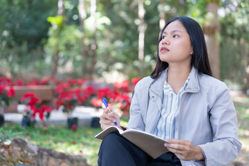 Attractive Asian female student sitting in the garden outside, taking notes, planning a business matter, looking upward thinking about something, concept work nomad lifestyle.