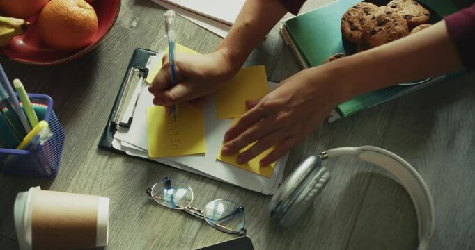 Close up a girl makes notes using a pen on yellow stickers word Hello and a joyful smiley face among items for studying work and snacking at home