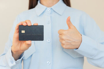 A young Caucasian woman dressed in a blue shirt holding a black bank card in her hand and giving a thumbs up.