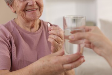 Caregiver giving glass of water to smiling senior woman with medical pill indoors, closeup