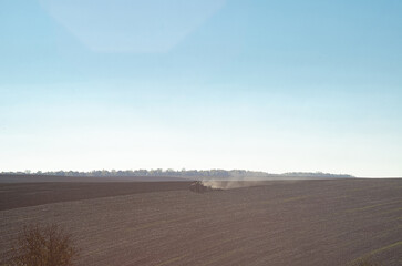 Obraz premium Agricultural spring view of field with black soil. Tractor plows in distance under blue sky, raising dust. Nostalgia.