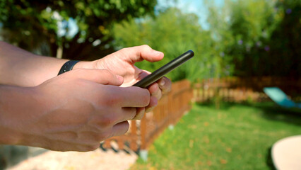 Person holding a smartphone in a bright backyard. Closeup of man hands holding a phone outdoors in a sunny garden. The natural light emphasize a relaxed, modern lifestyle in serene environment