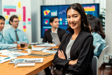 Portrait of happy young asian businesswoman or analyst looking at camera with her colleague analyzing data analysis in dynamic business strategy investment planning meeting. Habiliment