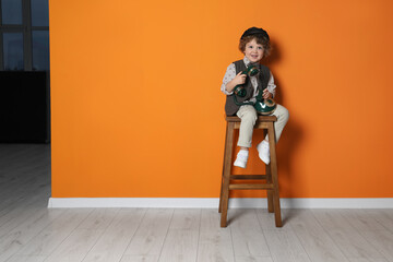 Cute little boy with old telephone on stool near orange wall, space for text