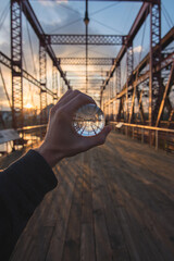 A perspective shot of a hand holding a glass sphere on a wooden bridge at sunset reflecting its structure within, creating a stunning backdrop of light, geometry, and artistic framing.