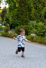 Child joyfully running in a green park. A young child in a playful shirt runs happily along a paved path in a lush park on a sunny day.