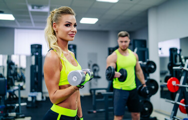 Dumbbell training enthusiasts. Two athletes train together in bright workout gear, focusing on strength with dumbbells at the gym.