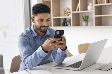 Young man sitting at workplace desk, holds smartphone, read sms, exchanging personal messages,...