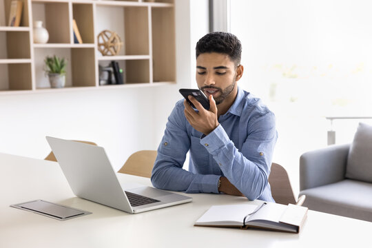 Young 20s man working seated at desk, holds smartphone lead talk on speakerphone, record message, send voice mail to colleague using messenger and mobile application, remote communication, modern tech
