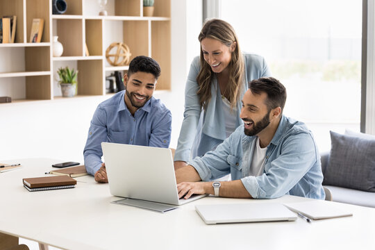 Three colleagues working together using laptop met in conference room in office, laughing while discuss collaborative task, learn new software, prepare online presentation. Cooperation, teamwork, tech