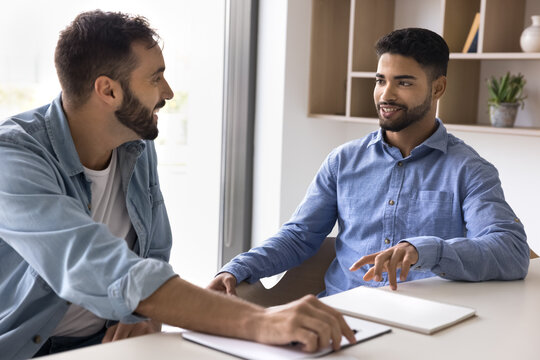 Multiethnic young teammates talking seated at table in modern office, smiling, collaborating on creative ideas and strategies, giving feedback on recent work, provide explanation, engaging in teamwork