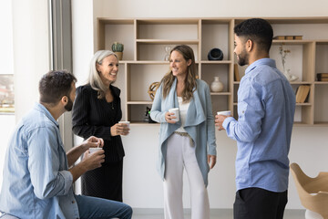 Group of four multiethnic young and older employees enjoy talk during coffee break in modern workplace, having fun, joking, looking satisfied with friendly, informal conversation met in office lobby