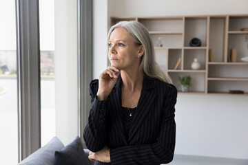 Professional 50s businesswoman in formal suit looking out window with pensive, thoughtful expression, ponder on upcoming project details, search issue solution, feels concerned standing in workspace