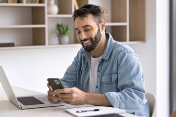 Smiling Latino man sit at workplace desk with laptop, distracted from online work, using modern smartphone, enjoy personal chat, check information, browse website, download new business application