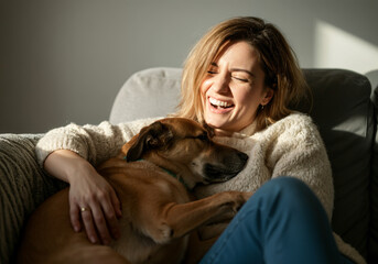 Happy woman embracing her dog while sitting on a comfortable sofa at home, enjoying a moment of affection and laughter
