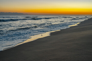 Gentle waves wash onto the dark sandy beach under a vibrant yellow and orange sunset sky.