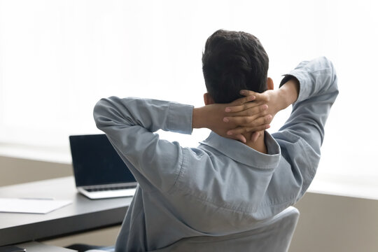 Taking pause. Rear back view tired young man sit at workplace stretch hands behind head relax from difficult workday take break for moment to rest procrastinate satisfied finishing task by end of day