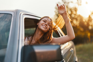 Happy young woman enjoying a road trip with her hair blowing in the wind, smiling from a car window...