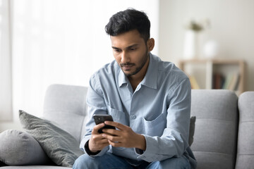 Concentrated young adult man freelancer work online from home sitting on sofa use cellphone spend...