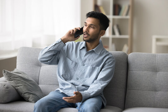 Making a phone call. Confident young male sitting on sofa speak on mobile telephone hold conversation chat discuss news with friend give remote consultation to client from home order delivery service