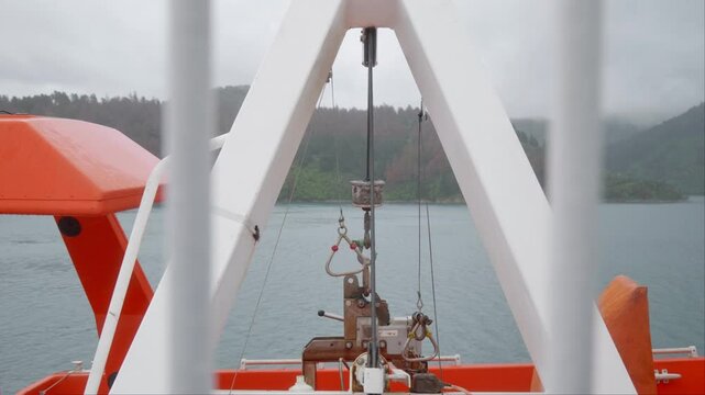 Close-up view of lifeboat davit hooks swaying gently in the wind above lifeboats on the portside of a ferry, with forested hills and overcast skies in the background