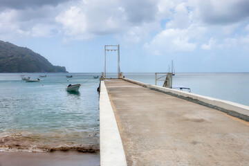 Obraz premium A long concrete pier stretching into the calm sea, with small fishing boats anchored nearby and a green hillside in the distance. Charlotteville, Tobago island, Caribbean sea. Copy space