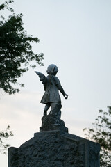 Fototapeta premium A weathered angel statue stands atop a stone pedestal, surrounded by trees. The figure, with delicate wings, captures a sense of timeless grace and quiet reflection against the backdrop of a clear sky