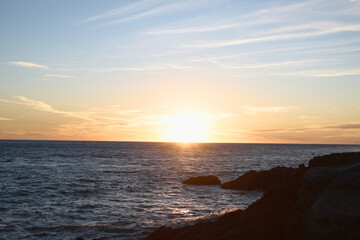 A serene ocean sunset with golden hues illuminating the horizon, soft waves reflecting the light, and rocky cliffs in the foreground under a pastel-colored sky. Peaceful and picturesque coastal scene.