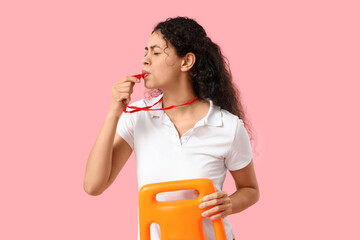 Beautiful young African-American female lifeguard with rescue buoy whistling on pink background