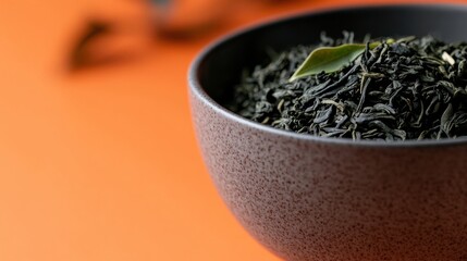 Gunpowder green tea leaves displayed in a textured bowl against a vibrant orange background in a serene setting