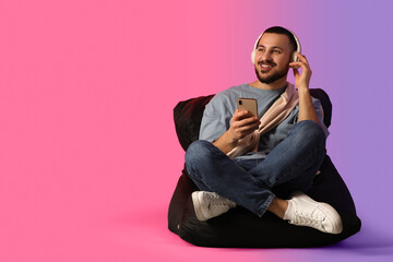 Young man in headphones with mobile phone on beanbag against color background