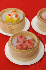 Steamed dumplings in bamboo baskets, featuring colorful fillings, presented on white plates against a vibrant red backdrop.
