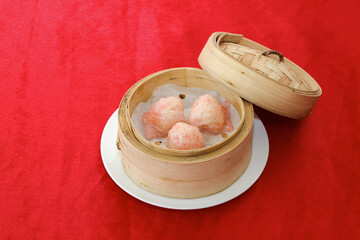 A bamboo steamer containing three pink dumplings sits on a white plate against a vibrant red background.