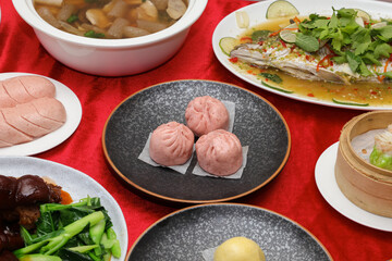 A vibrant spread of Asian cuisine featuring dumplings, fish, vegetables, and a rich soup, set against a red tablecloth.