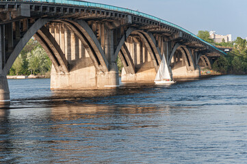 Obraz premium A serene view of a sailboat gliding under the arches of a Kyiv bridge, with sunlight casting warm reflections on the water, capturing the harmony of architecture and nature.