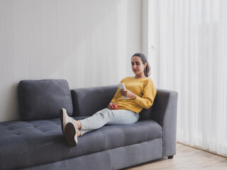 Woman latin american one person smile happy relax comfortable sitting sofa and look hand holding use remote control adjust air conditioning inside on most wonderful vacation day in living room house.