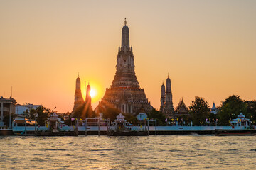 Fototapeta premium As the sun sets behind Wat Arun, its ornate spires glow with warm golden light, casting beautiful reflections on the tranquil waters of the Chao Phraya River. A serene moment in Bangkok.