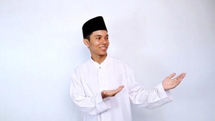Handsome young Asian Muslim man smiling showing his hand recommending something on the side isolated on white background