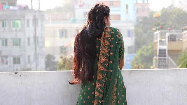 A Bengali woman, wearing a beautiful green block-printed salwar kameez, stands alone on a rooftop in the afternoon. The air moves her long black hair and clothes. She looks outside, lost in thought.