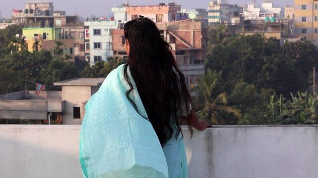 A Bengali woman, clad in a blue cotton saree, stands alone on a rooftop in the afternoon, enjoying the fresh air as it blows around her. She gazes out at the view.