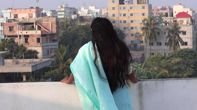 A Bengali woman, clad in a blue cotton saree, stands on the rooftop, enjoying the cool breeze. She gazes out at the scenery, her back to the viewer.