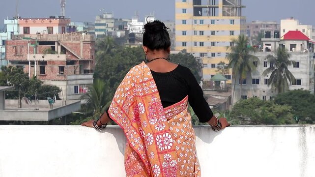 A Bengali woman in a beautiful floral cotton saree stands alone on a rooftop on a sunny day, looking out at the outdoor environment. Back view.