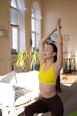 Calm Asian female in sportswear practicing yoga, breathing deeply, raising hands and meditating in...