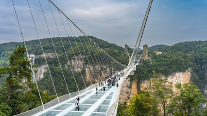 The longest glass suspension bridge in the world. People walk over a precipice on a transparent surface. Stretched support cables against the sky and rocks. China. Zhangjiajie Glass Bridge