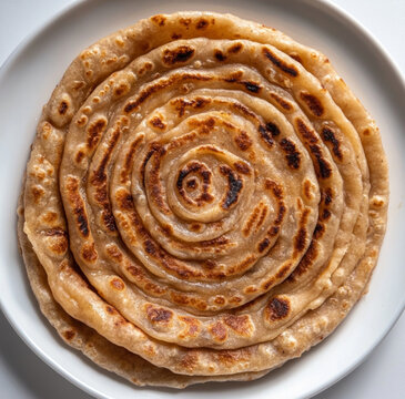 A freshly made whole wheat Lachha Paratha with perfectly crisp, brown layers, white plate. The paratha has a rich, flaky texture with visible layers spiraling outwards,the plate is placed on a neutral
