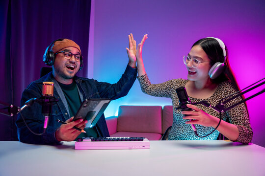 Young asian man and woman host streaming podcast with condenser microphone work on tablet at home broadcast studio. Content creator blogger recording voice over radio interview guest conversation - Powered by Adobe