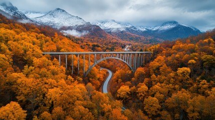 Stunning Aerial View of a Modern Bridge Spanning Fall-Foliage Covered Landscape under Dramatic Sky with Snow-Capped Mountains in the Distance