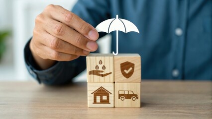A person places an umbrella atop wooden blocks displaying icons representing insurance, protection, and various assets like home and car.